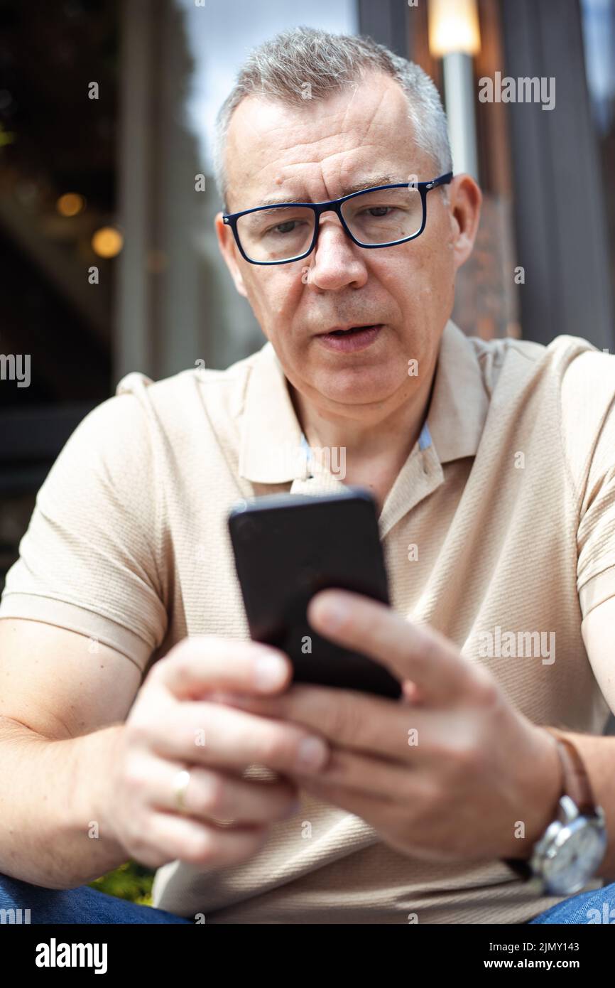 Homme d'affaires senior aux cheveux gris assis dans des lunettes à l'extérieur et à envoyer des SMS sur un smartphone. Style de vie moderne, communication sociale, processus de travail Banque D'Images