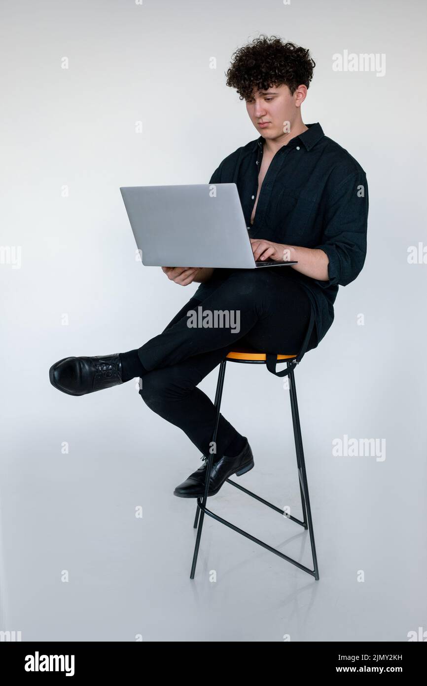 Photo pleine longueur de mécontent jeune homme en forme de curly dans l'aspect noir total assis à pieds croisés sur la chaise et de jouer à des jeux d'ordinateur sur ordinateur portable moderne, studio gris Banque D'Images