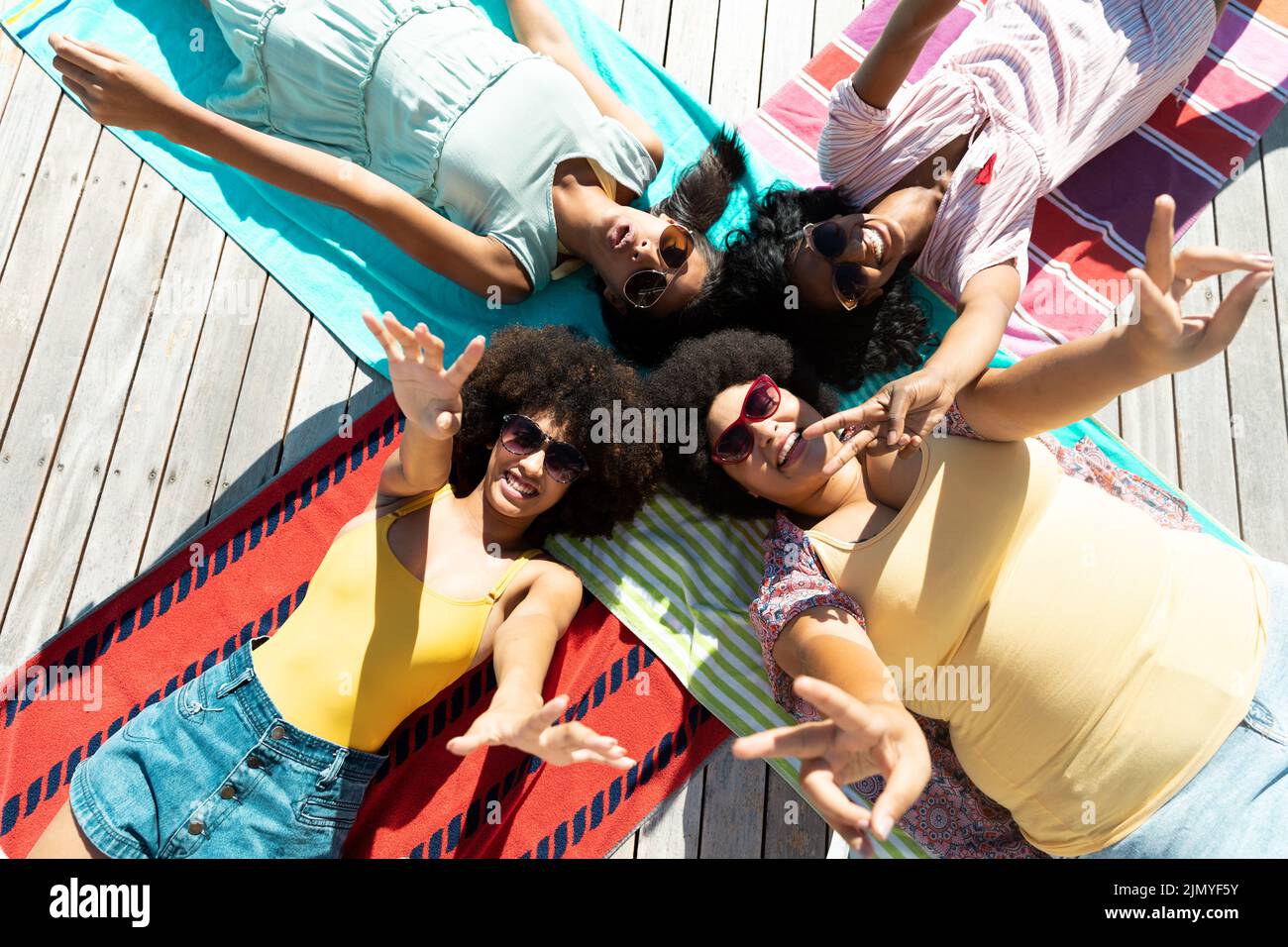 Vue en grand angle des amis biraciaux heureux portant des lunettes de soleil faisant des gestes tout en étant allongé sur des serviettes au bord de la piscine Banque D'Images