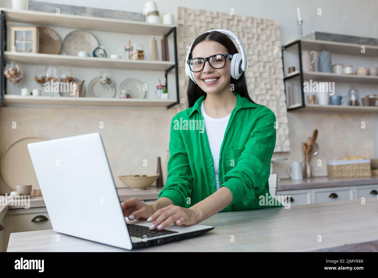 Portrait d'une jeune étudiante du millénaire étudiant en ligne à la maison à l'aide d'un ordinateur portable portant un casque, des vêtements verts et des lunettes. Il regarde la caméra, sourit. Banque D'Images