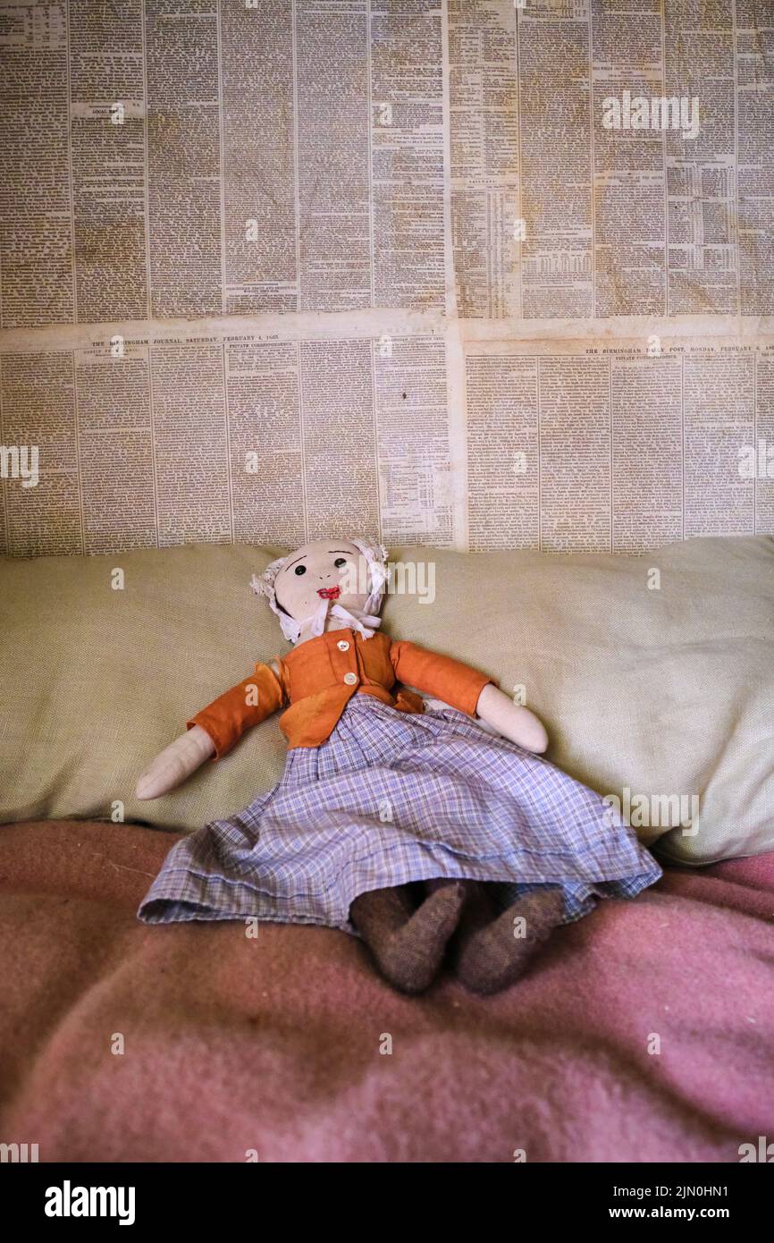 Détail d'un jouet de poupée de chiffon d'enfant typique dans une robe, jupe, sur un lit. Papier peint du journal. Dans une récréation d'une maison de travail typique, générique, maison Banque D'Images