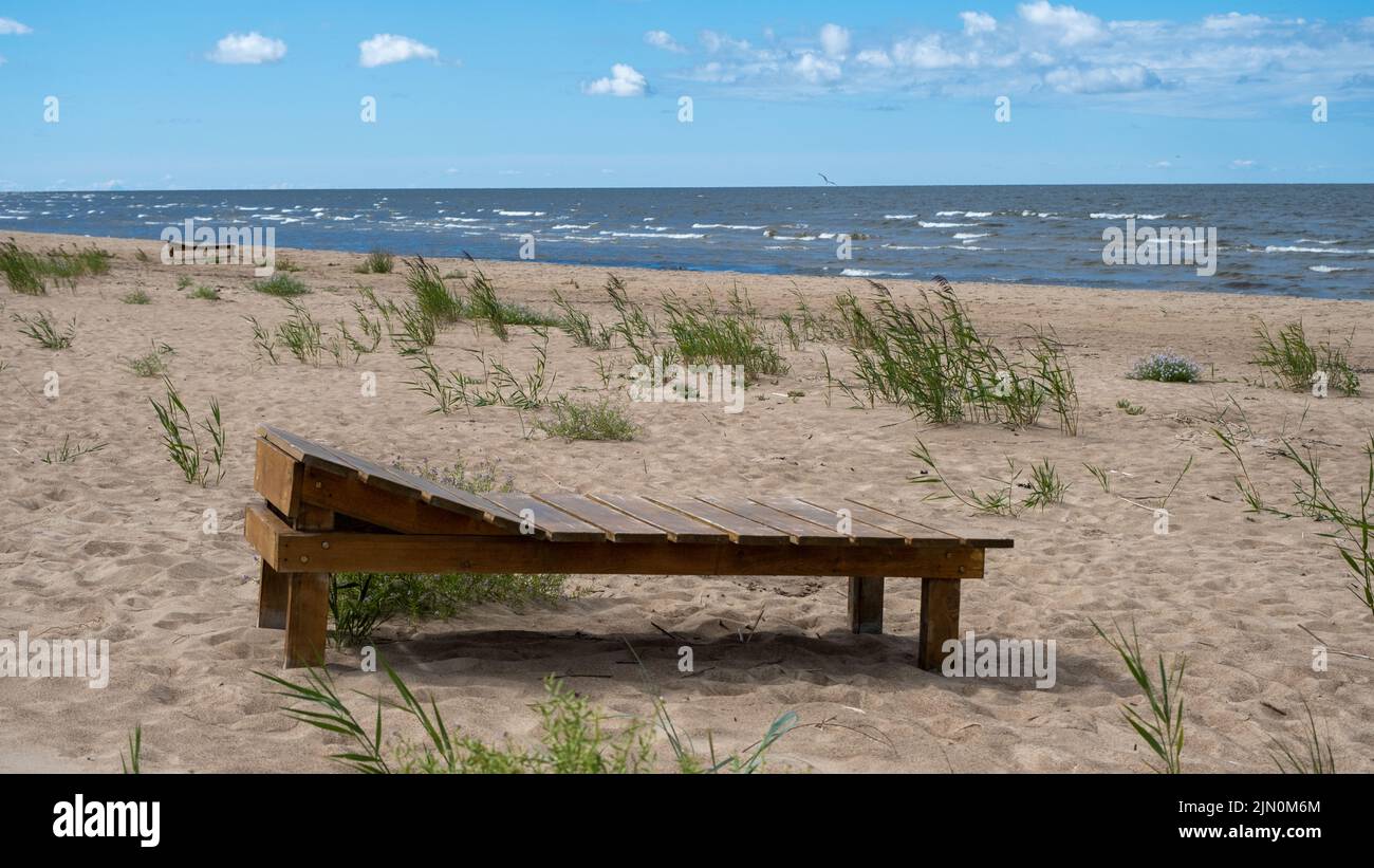 Un lit de plage en bois marron reposant sur une plage de sable au bord de la mer Baltique dans le golfe de Riga, des vagues en arrière-plan et le soleil chaud de midi coulent Banque D'Images