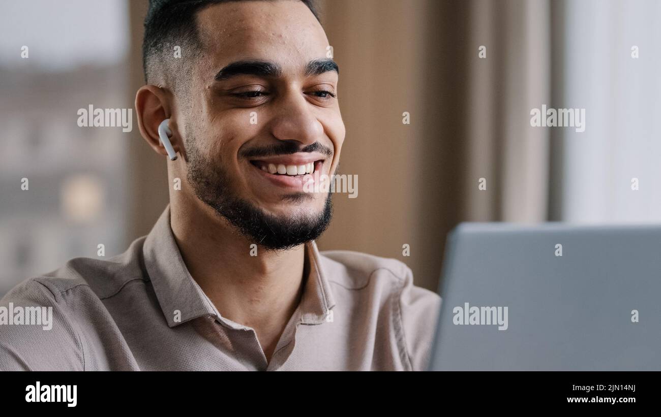 Joyeux jeune arabe homme porter des écouteurs sans fil regarder l'ordinateur portable se détendre regarder la comédie vidéo drôle film sourire profiter de pause libre lire les bonnes nouvelles Banque D'Images