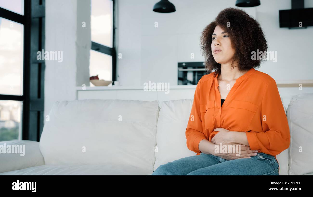 Triste femme africaine à la maison cheveux bouclés fille malade assise sur un canapé dans la cuisine salon tenant le ventre touchant l'abdomen souffrant de maux d'estomac douloureux Banque D'Images