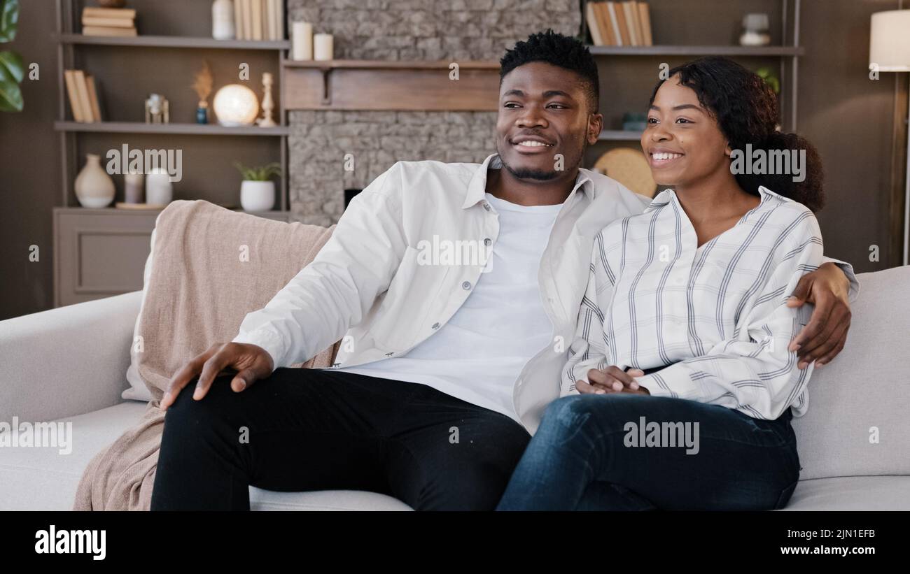 Un couple heureux et récemment marié, un homme et une femme africains assis à la maison, regardant la télévision, parlant rire. Mari et femme discutent avec un thérapeute de famille Banque D'Images