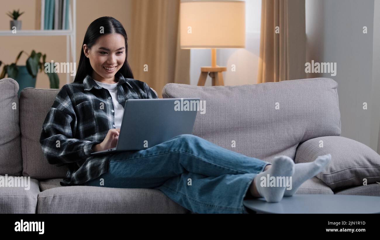 Femme asiatique souriante utilisant ordinateur portable navigation décontractée recherche en ligne jouer jeu d'ordinateur. Une jeune fille étudiante coréenne indépendante assise sur un canapé e-learning Banque D'Images