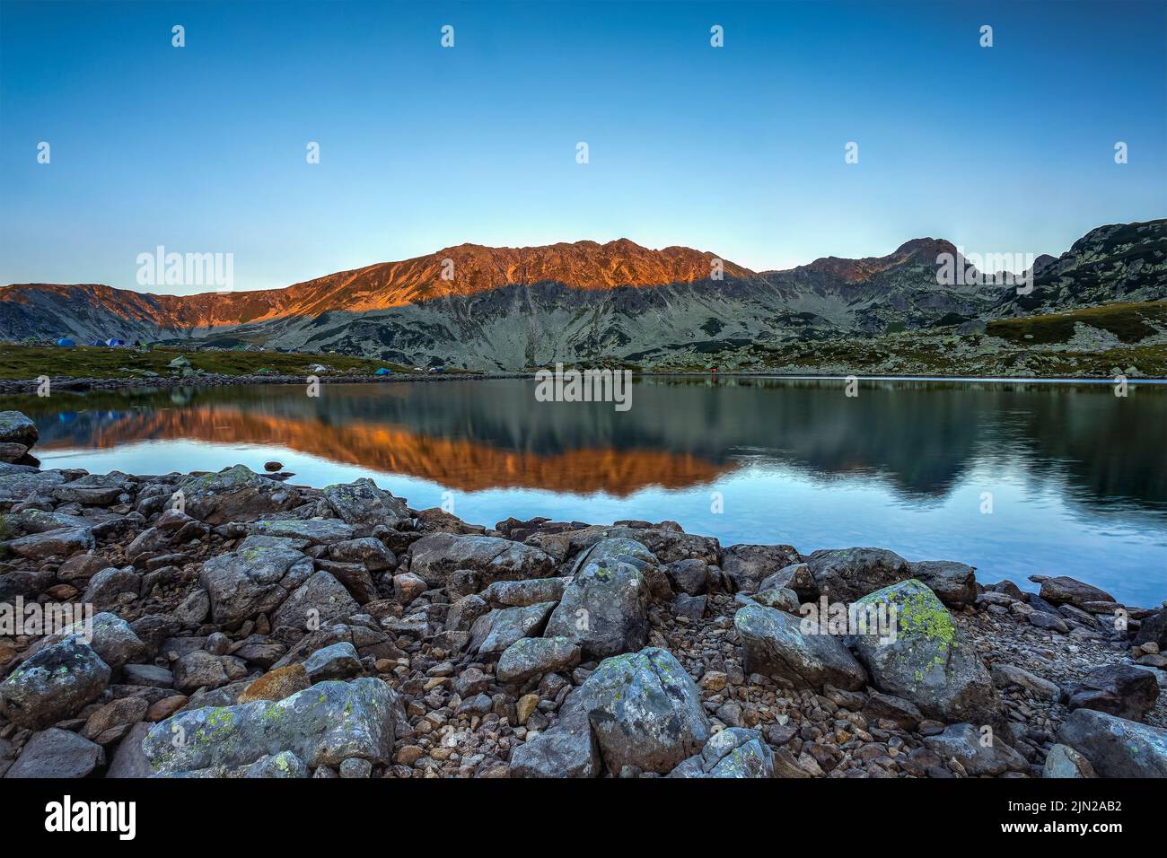 Lever de soleil avec lumière douce et orange illuminant les sommets des montagnes de Retezat et se reflétant dans le lac Bucura. Photo prise le 7th août 2022 sur la rive Banque D'Images