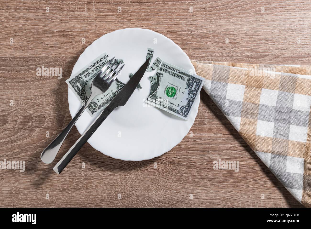 Une assiette blanche avec une facture de dollar déchirée en deux grands morceaux et quelques petits quartiers. Sur la plaque se trouve un couteau et une fourchette. Il est sur une table en bois et ne Banque D'Images