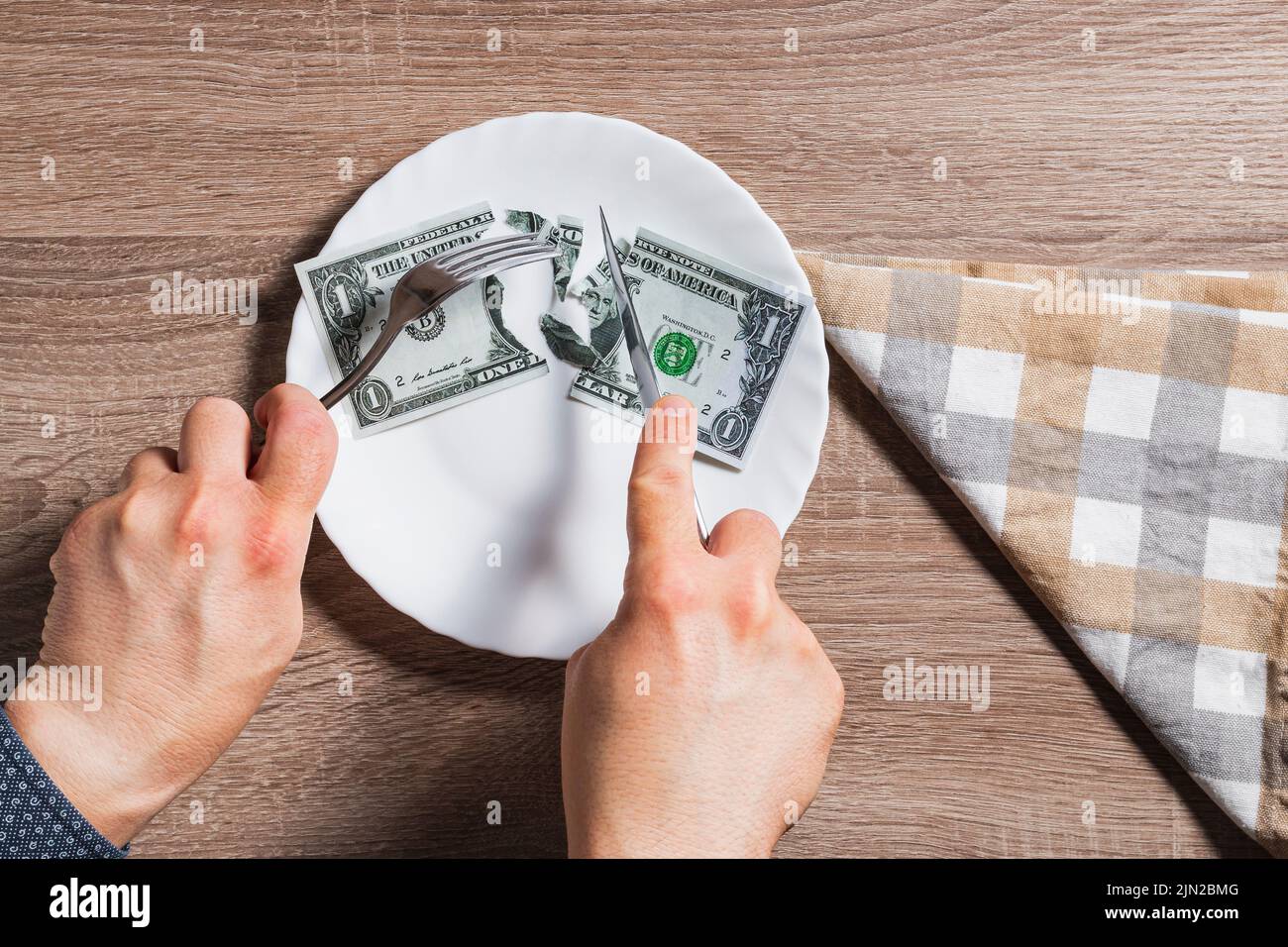Mains d'une personne inconnue coupant une facture de dollar dans une assiette avec couteau et fourchette. Il est sur une table en bois et à côté il y a une serviette en tissu. Banque D'Images