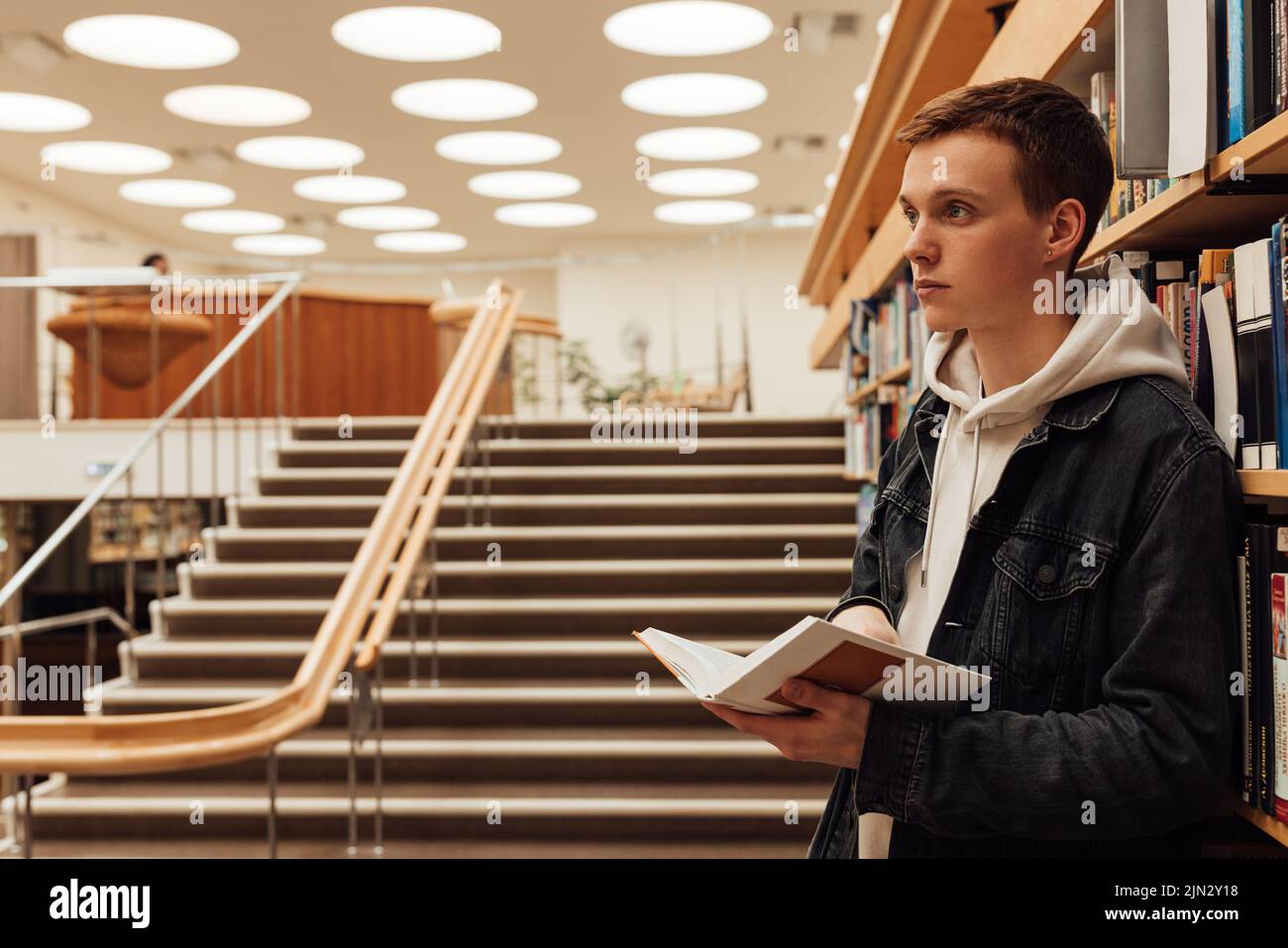 Vue latérale d'un étudiant de sexe masculin dans la bibliothèque contenant un livre. Étudiant de l'université debout à la bibliothèque. Banque D'Images