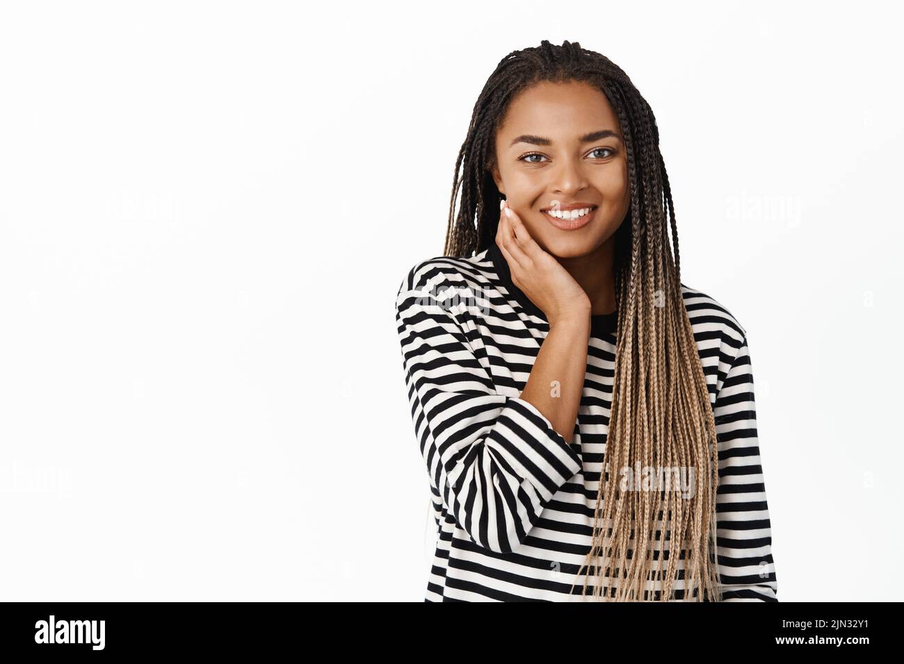 Belle jeune fille noire avec peau naturelle du visage sans maquillage, touchant son visage et souriant avec plaisir, debout sur fond blanc Banque D'Images