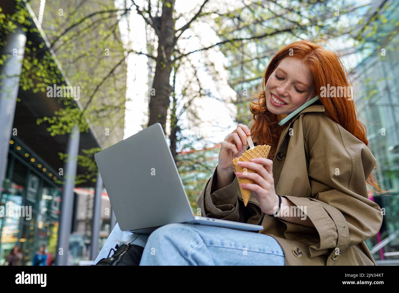 Ado redhead hipster fille utilisant un ordinateur portable parlant sur le téléphone dans la rue urbaine de ville. Banque D'Images