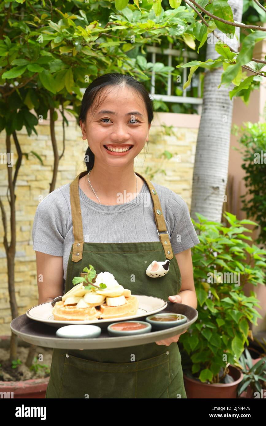 Une serveuse vietnamienne sert des gaufres belges avec des fruits et de la crème glacée dans un café extérieur Banque D'Images
