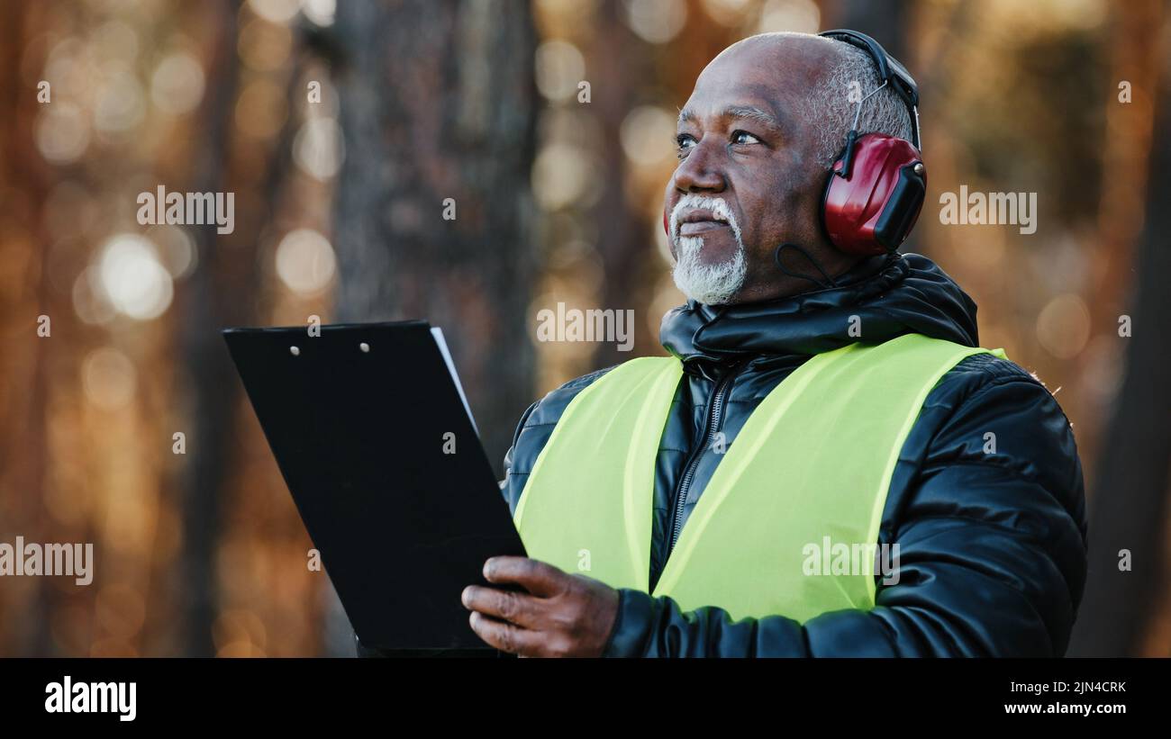 Gros plan afro-américain homme contremaître professionnel ingénieur forestier debout à l'extérieur, portant des écouteurs de protection pour évaluer la situation dans la fabrication de l'arbre Banque D'Images