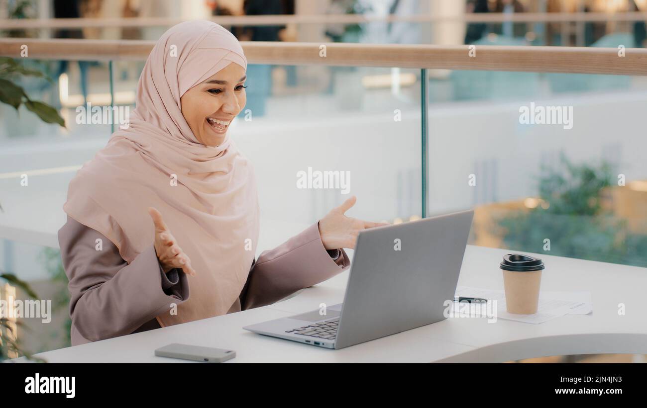 Bonne jeune femme d'affaires arabe assise au bureau, dactylographiant sur un ordinateur portable qui travaille sur Internet, vérifiant une nouvelle application souriant en regardant l'appareil photo avec le pouce vers le haut Banque D'Images