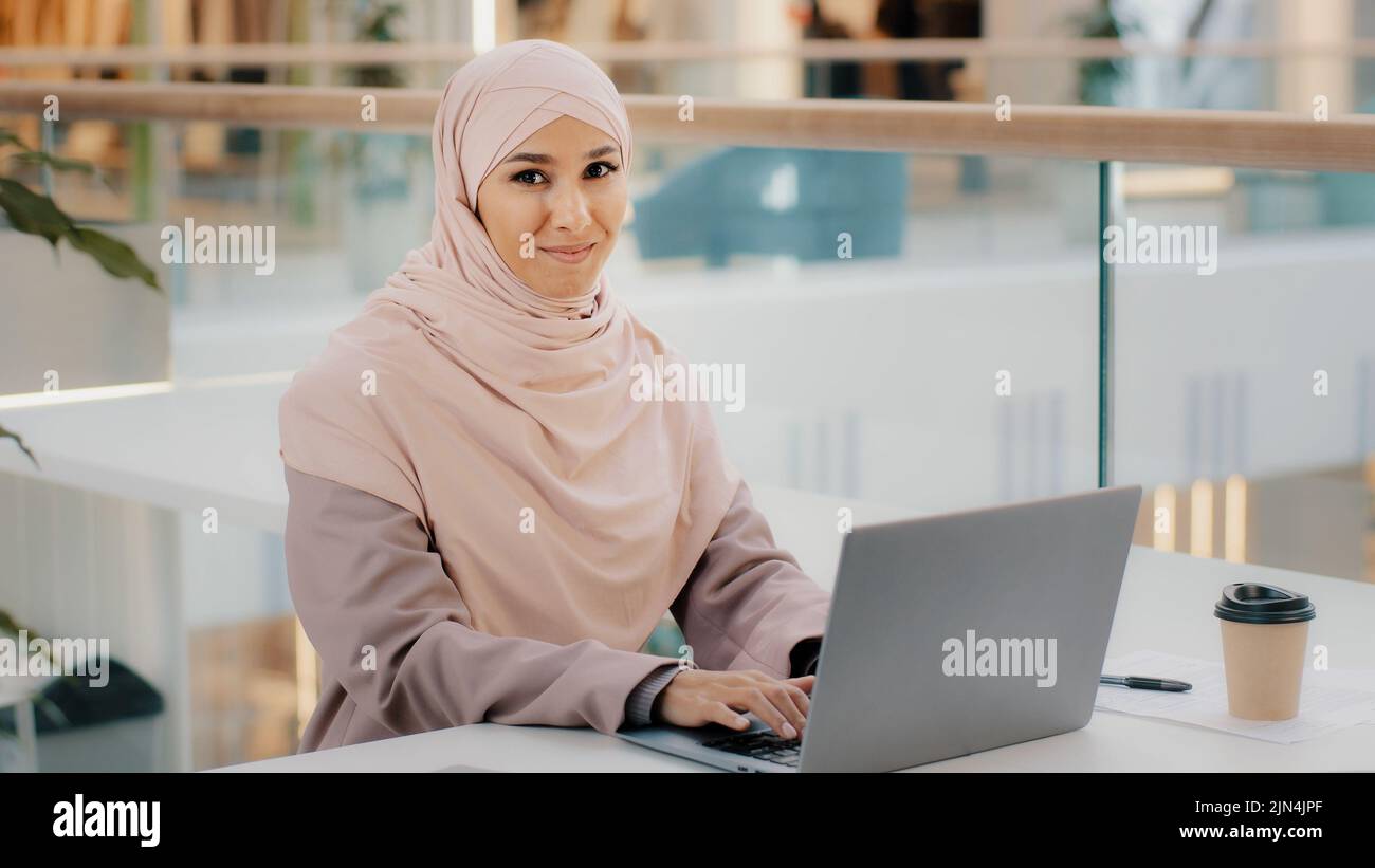 Bonne jeune femme d'affaires arabe assise au bureau, dactylographiant sur un ordinateur portable qui travaille sur Internet, vérifiant une nouvelle application souriant en regardant l'appareil photo avec le pouce vers le haut Banque D'Images