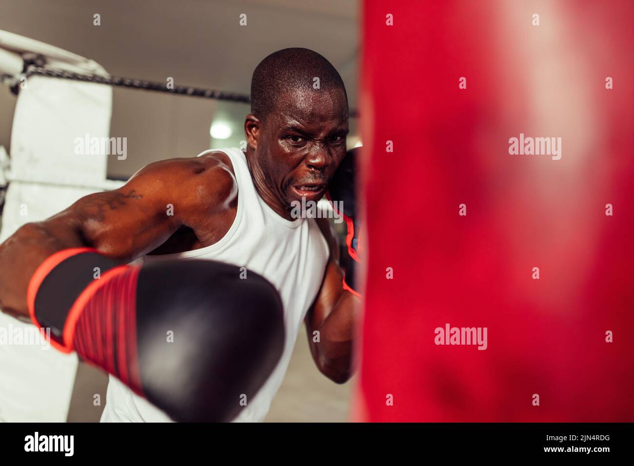 Un jeune combattant balançant un poing ganté dans un sac de poinçonnage rouge dans une salle de sport. Sueur jeune homme ayant une séance d'entraînement dans un gymnase de boxe. Banque D'Images