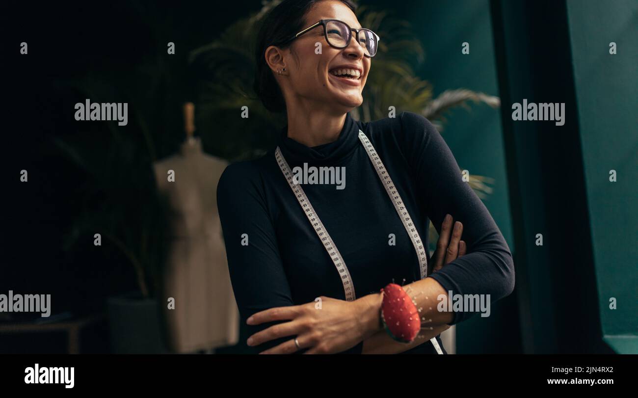 Riant femme designer de mode dans son studio. Femme asiatique debout avec du ruban de mesure autour du cou et regardant loin. Banque D'Images