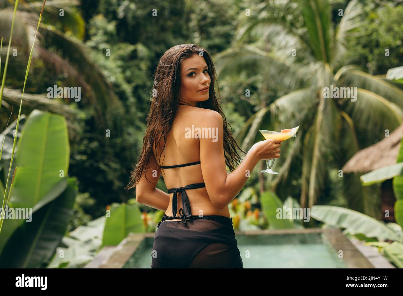 Photo en plein air d'une jeune femme avec un cocktail au bord de la piscine, elle porte un bikini noir et regarde l'appareil photo. Banque D'Images
