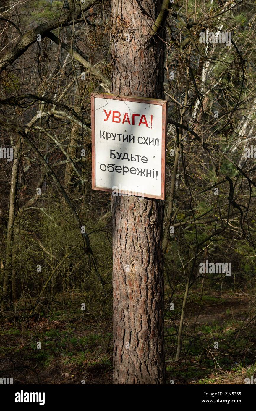 Panneau d'affichage blanc avec texte d'avertissement sur le tronc de pin dans la forêt de printemps ensoleillée. Traduction de l'ukrainien: Avertissement! Une pente raide soyez prudent ! Banque D'Images