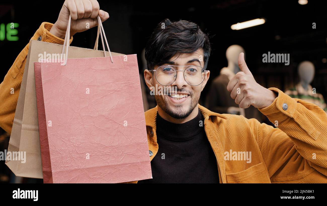 Jeune beau gars joyeux se tient dans la boutique tenant des sacs de papier points pour bon achat heureux homme élégant dans le magasin de vêtements montrant le pouce vers le haut geste Banque D'Images