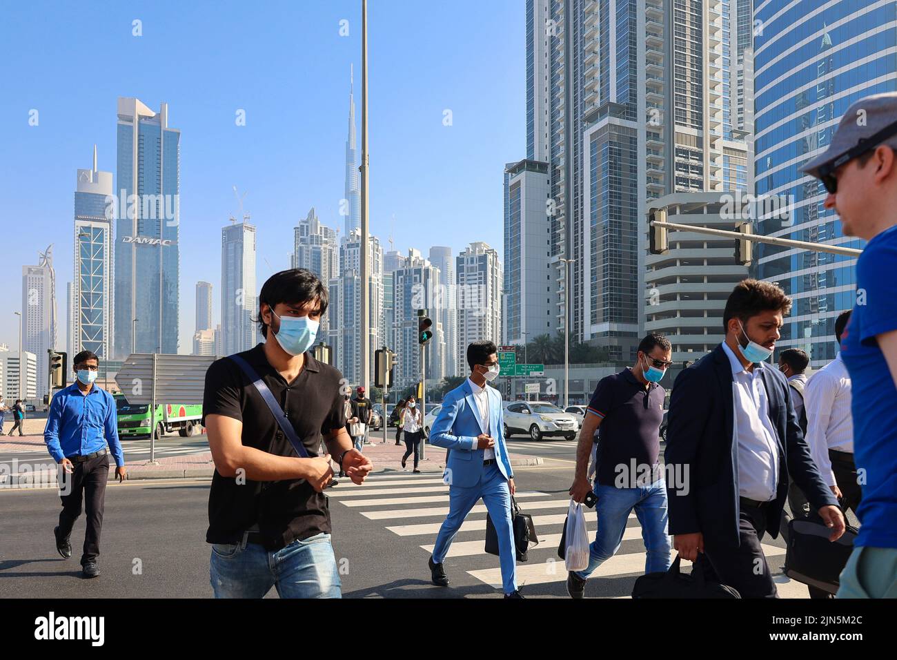 Eau, Dubaï - 27 novembre 2021 : les gens marchent le long de la traversée piétonne dans le centre d'affaires de Dubaï. Les hommes en costume et les masques médicaux se précipitent pour travailler sur fond d'énormes gratte-ciel. Photo de haute qualité Banque D'Images