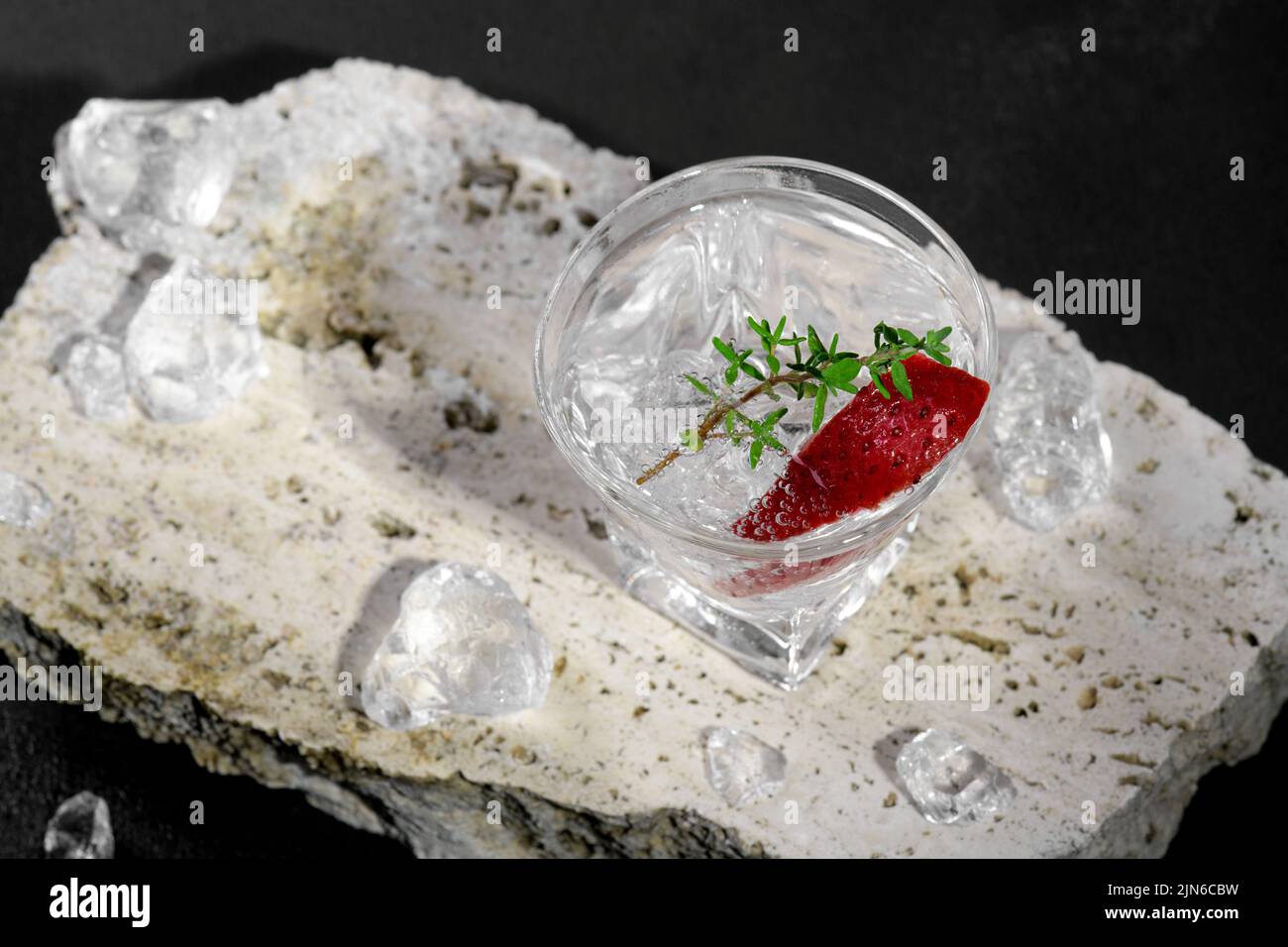 Vodka en verre à glace, décorée de tranches de fraises et de thym sur le podium de travertin sur fond de pierre sombre. Boisson alcoolisée avec stra Banque D'Images