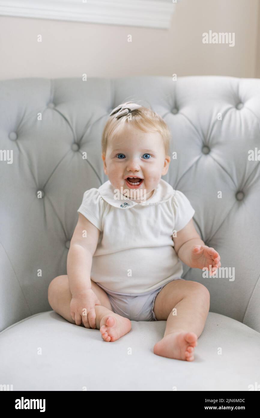 Bébé fille s'assoit dans un fauteuil gris et sourit Banque D'Images