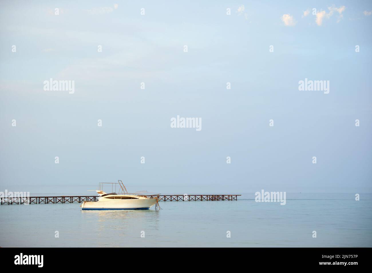 Le bateau à moteur blanc flotte dans l'eau de l'océan sur le long pont de la jetée sous le ciel bleu clair, vue depuis la plage de sable. Concept de voyage d'été Banque D'Images