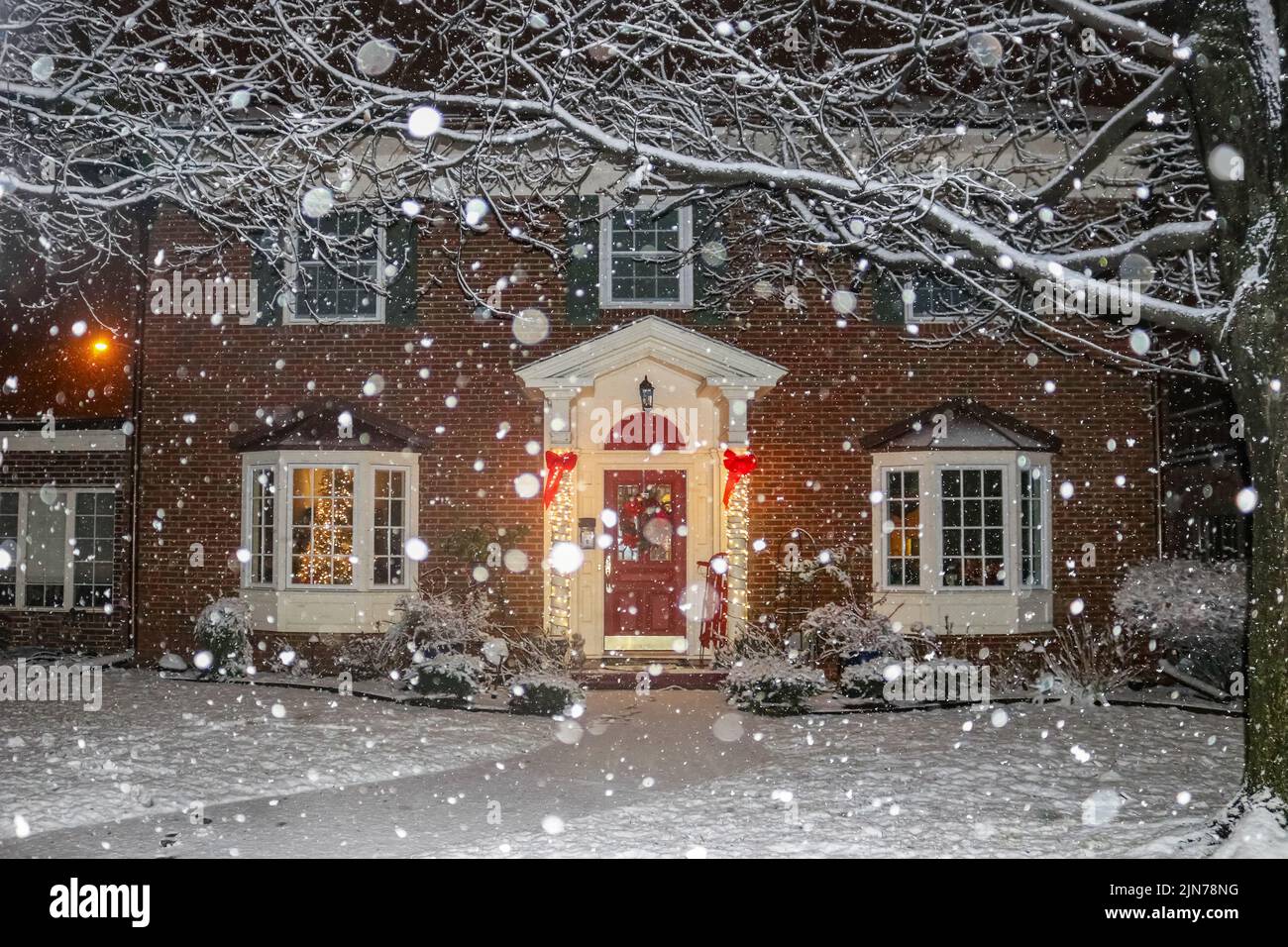 Chute de neige sur une belle maison en briques avec colonnes et baies vitrées avec arbre de Noël illuminé et traîneau rouge et couronne sur le porche Banque D'Images