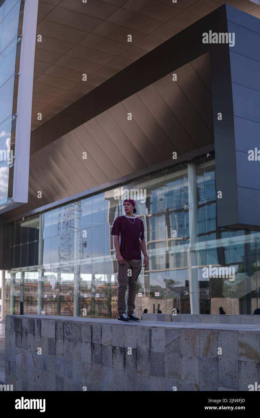 Latino jeune homme habillé de vêtements urbains posant sur une grande clôture avec l'architecture en arrière-plan. Un homme urbain hispanique qui se tient stoïquement devant lui Banque D'Images