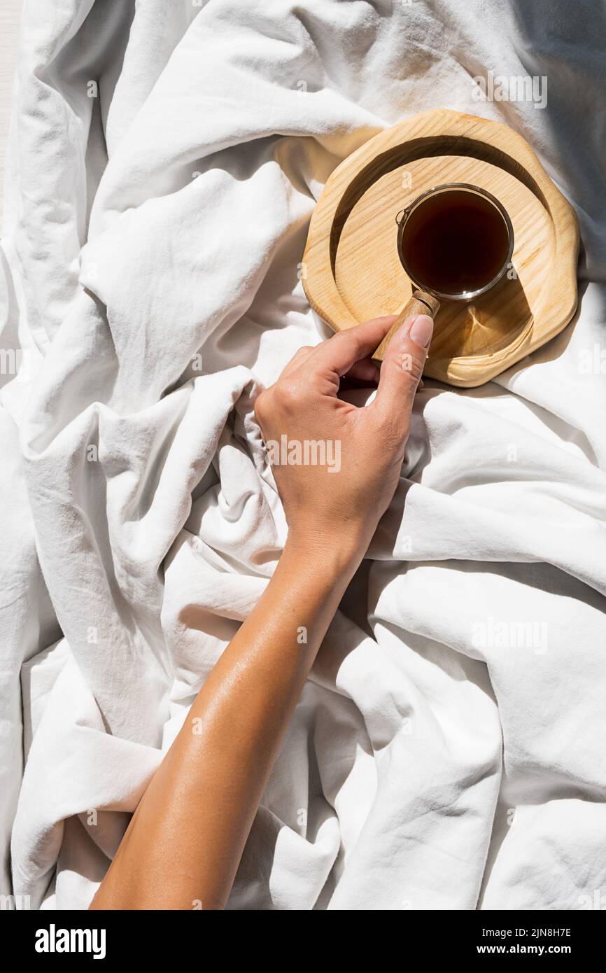 Les femelles atteignent la main pour une tasse de café du matin dans le lit. Une femme se réveille à la lumière du soleil sur une literie blanche. Banque D'Images