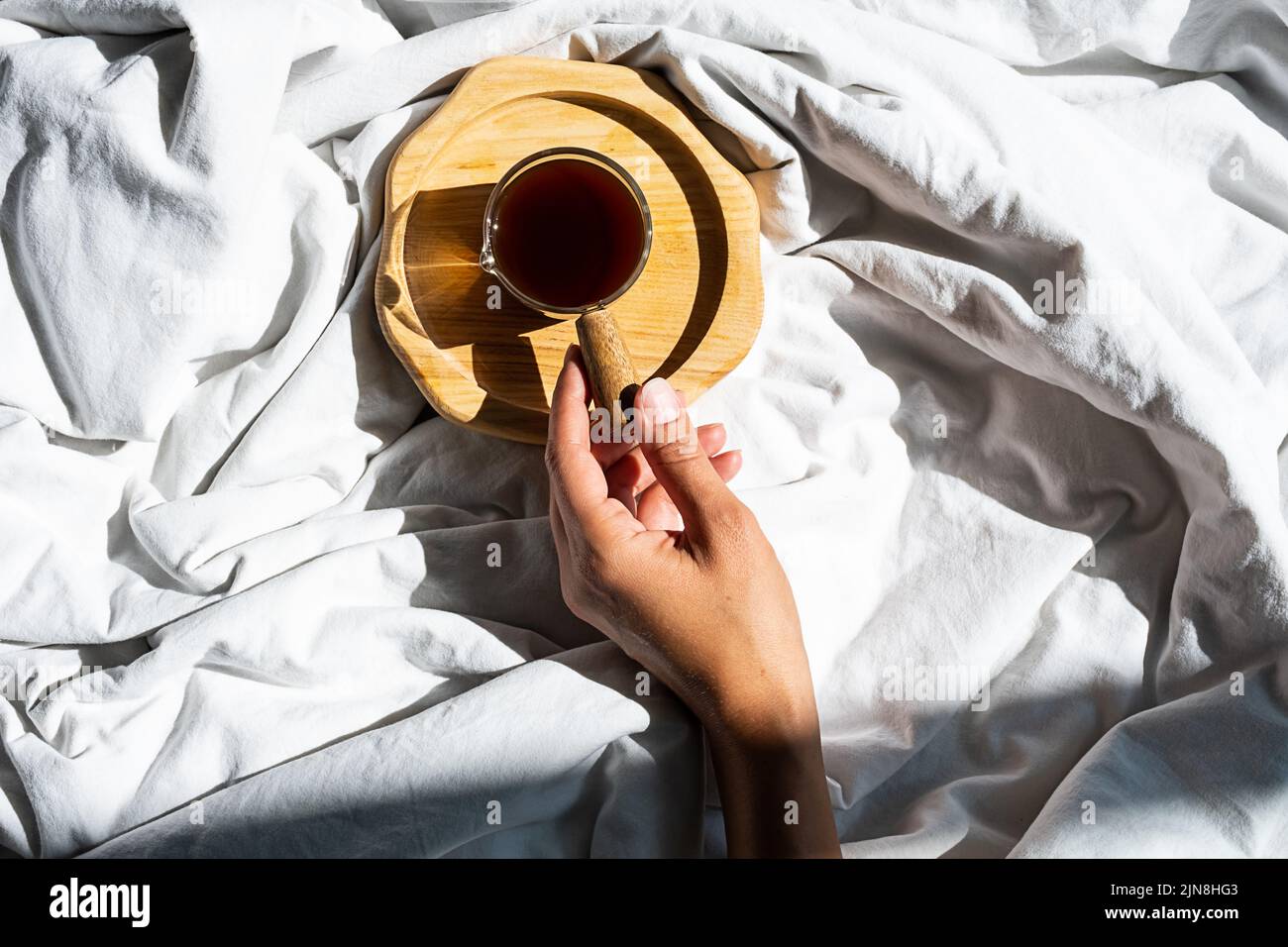 Les femelles atteignent la main pour une tasse de café du matin dans le lit. Une femme se réveille à la lumière du soleil sur une literie blanche. Banque D'Images