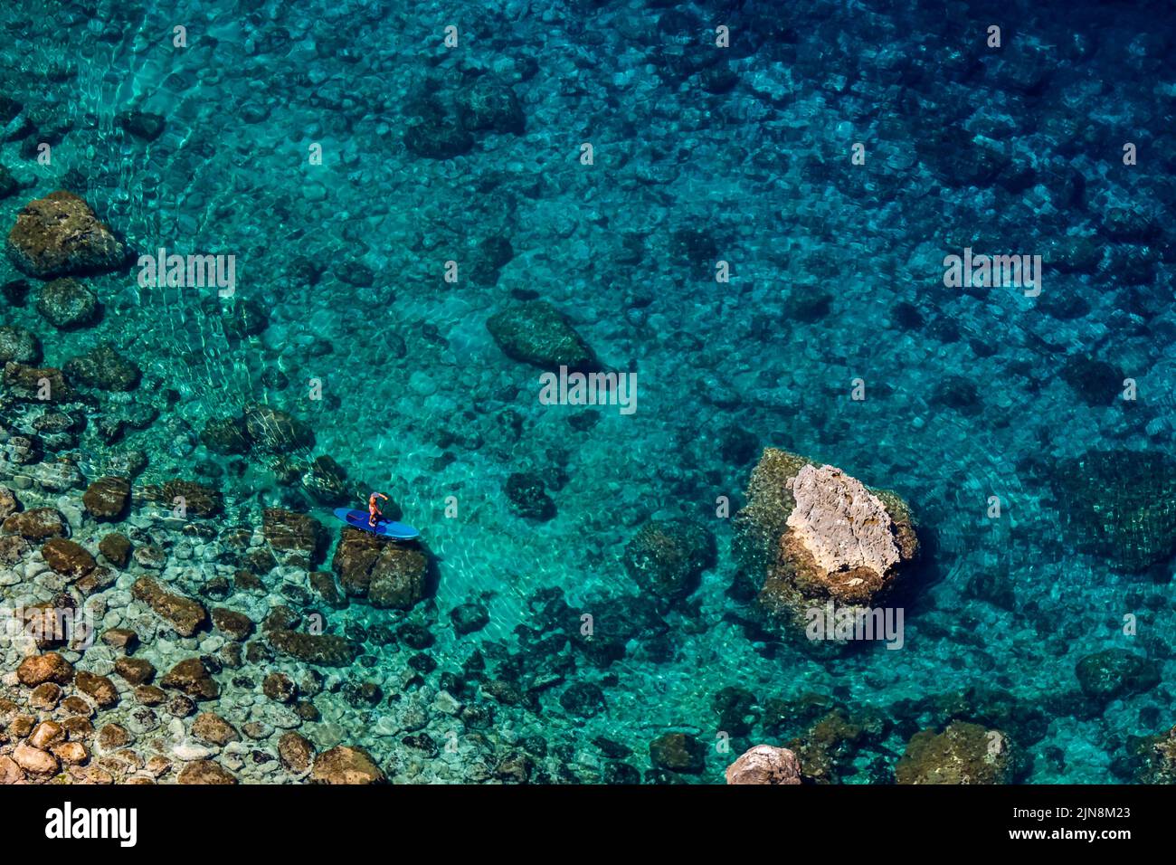 Photographie du dessus d'un adulte méconnaissable debout sur un panneau bleu SUP Stand Up, entre les rochers sur la rive turquoise de l'eau. Banque D'Images