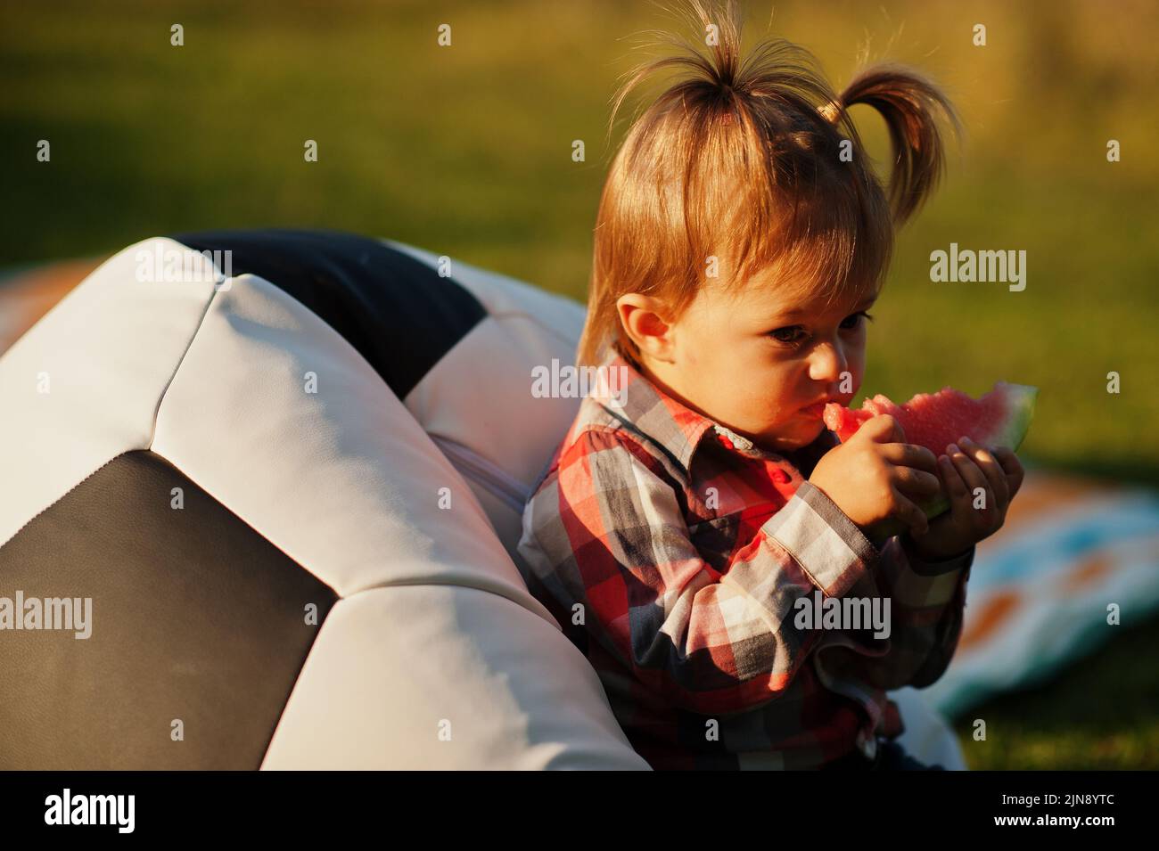 Petite fille mignonne en chemise à carreaux, asseyez-vous sur un pouf de ballon de football et mangez de la pastèque. Banque D'Images