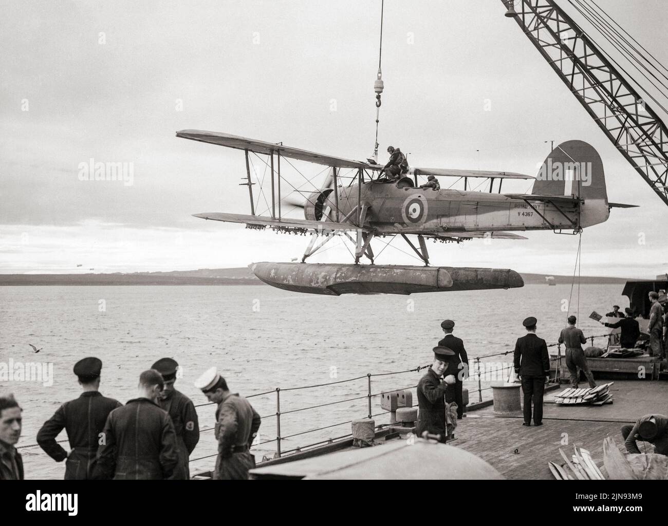 Après un vol de reconnaissance, un avion marin Fairey Swordfish retourne au HMS Malaya et est hissé à bord. Bien que obsolète en 1939, l'espadon a remporté quelques succès spectaculaires pendant la guerre. Parmi les événements notables, citons la célèbre attaque contre le cuirassé allemand Bismarck, qui a contribué à sa disparition. L'espadon a coulé un plus grand tonnage de transport de l'axe que tout autre avion allié pendant la guerre et est resté en service de première ligne jusqu'au jour V-E, après avoir survécu à une partie de l'avion destiné au remplacer. Banque D'Images