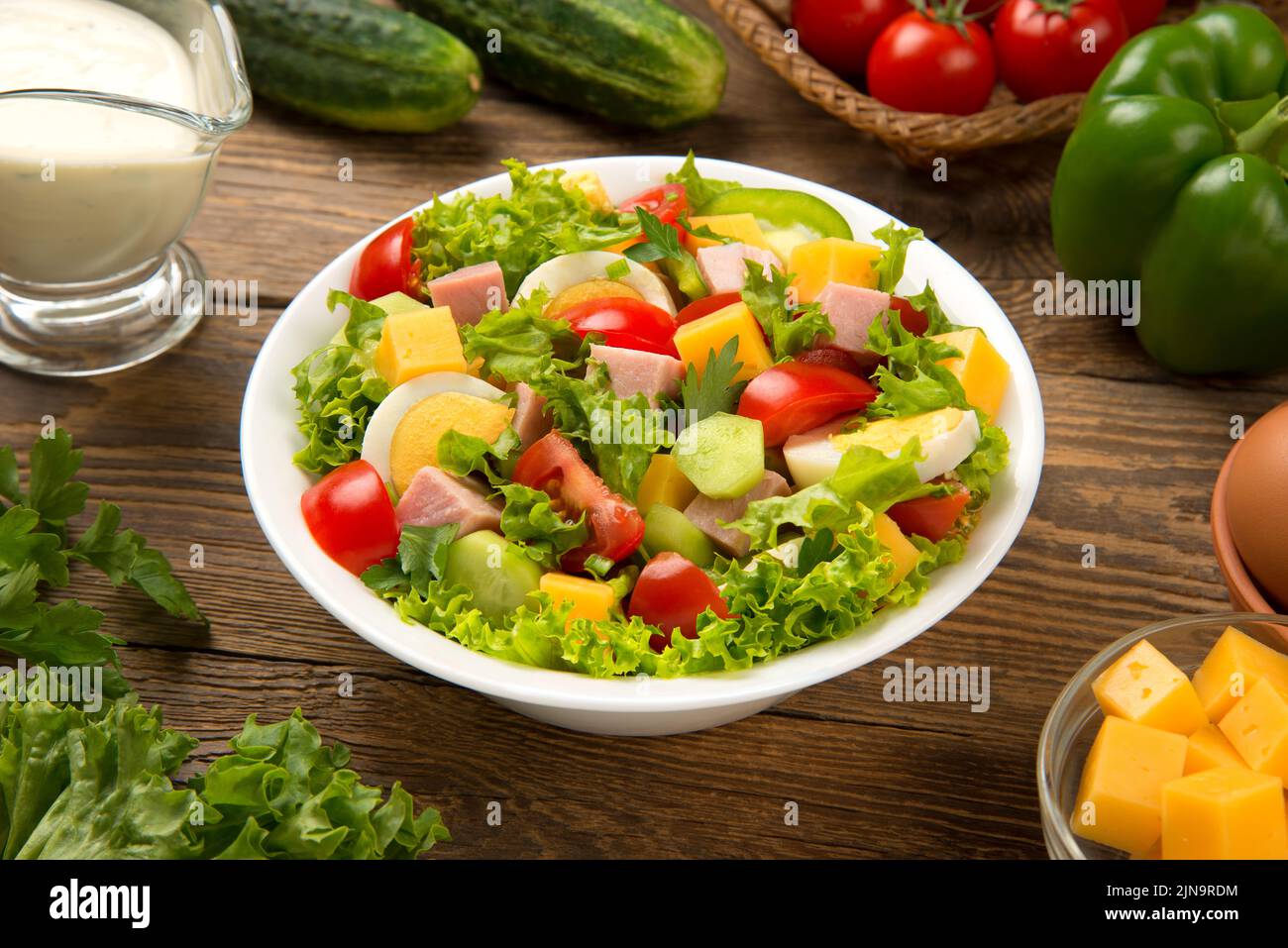 Salade du chef avec tomate, concombre, laitue, jambon, poivron, fromage et œufs dans un saladier blanc sur une table rustique en bois. Mise au point sélectionnée. Banque D'Images