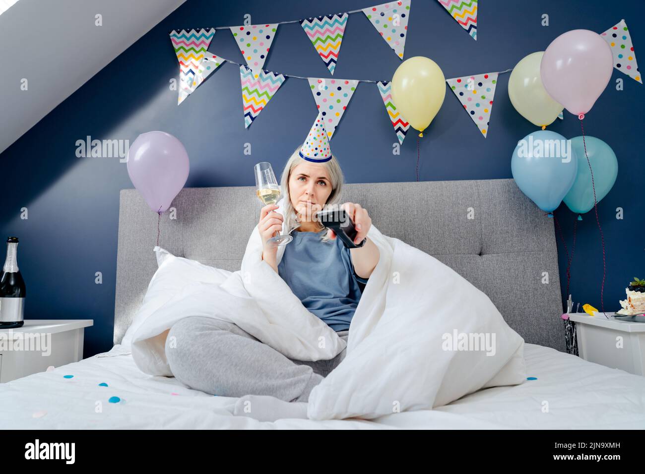 Une femme triste en pyjama, un chapeau de fête boit du champagne et regarde la télévision, enveloppée dans une couverture tout en étant assise sur un lit dans une chambre décorée. Célèbre un birt Banque D'Images Une femme triste en pyjama, un chapeau de fête boit du champagne et regarde la télévision, enveloppée dans une couverture tout en étant assise sur un lit dans une chambre décorée. Célèbre un birt Banque D'Images