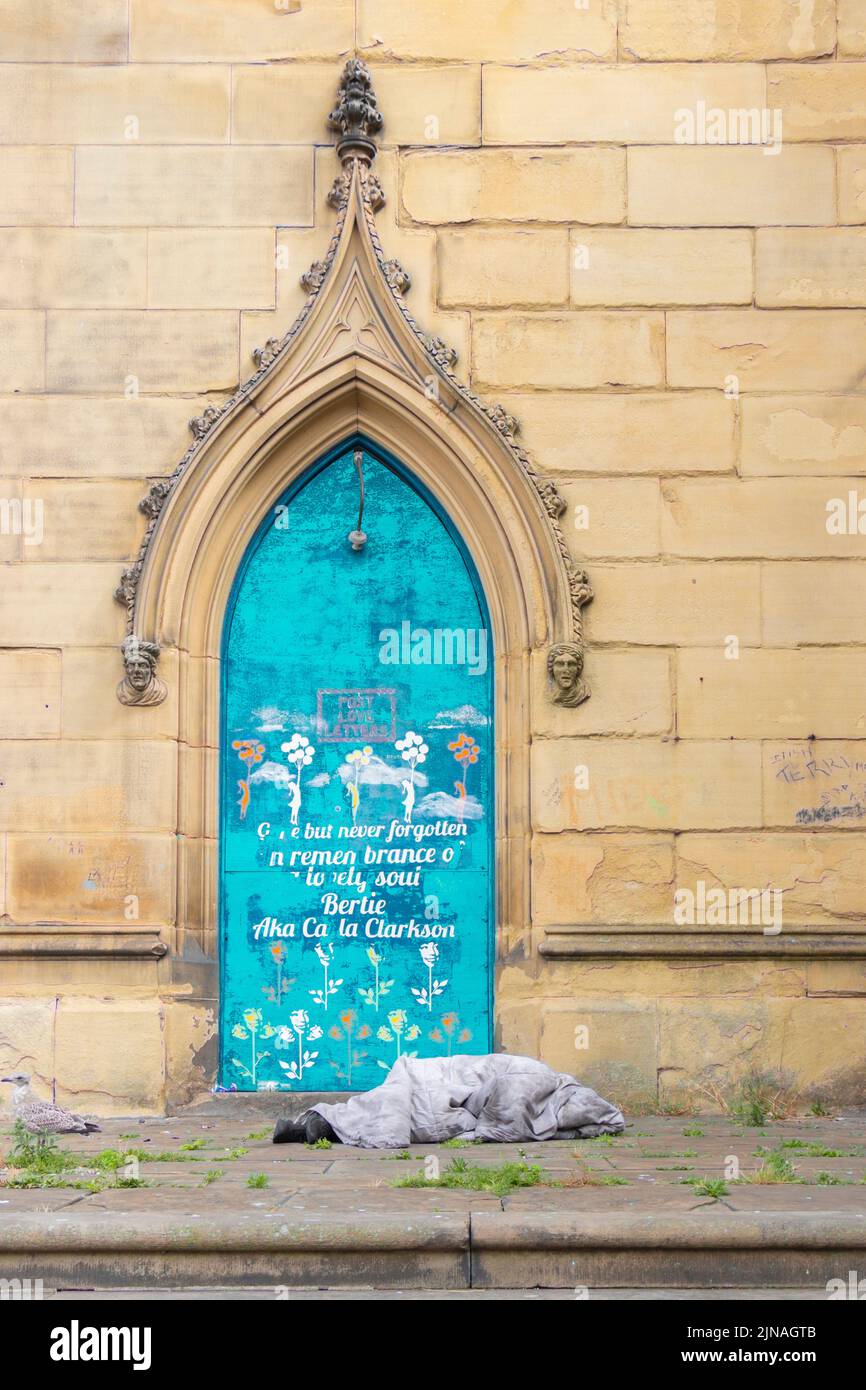 Un sans-abri dort sous une porte arquée bleue au design complexe, sertie dans un grand mur de pierre avec une citation gravée. disparu mais jamais oublié Banque D'Images