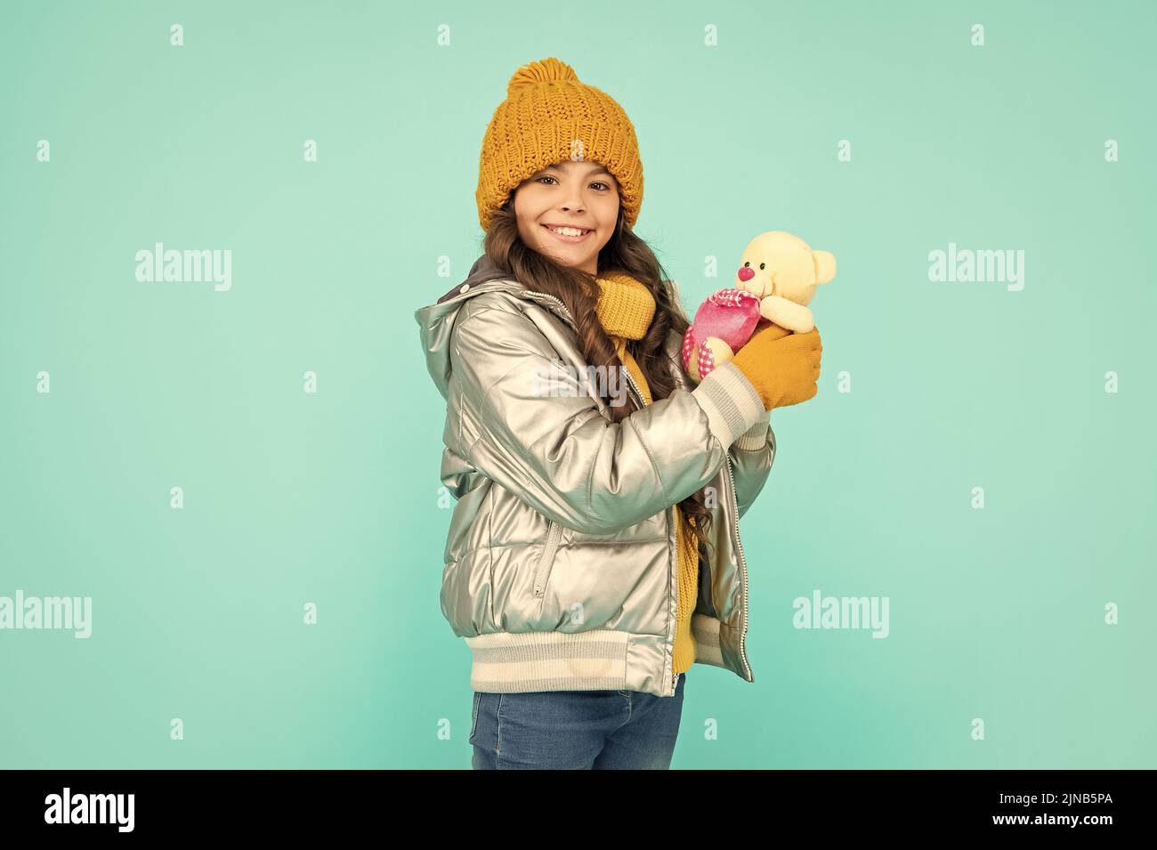 une jeune fille souriante tient un ours en peluche. enfant portant des vêtements chauds sur fond bleu. Banque D'Images