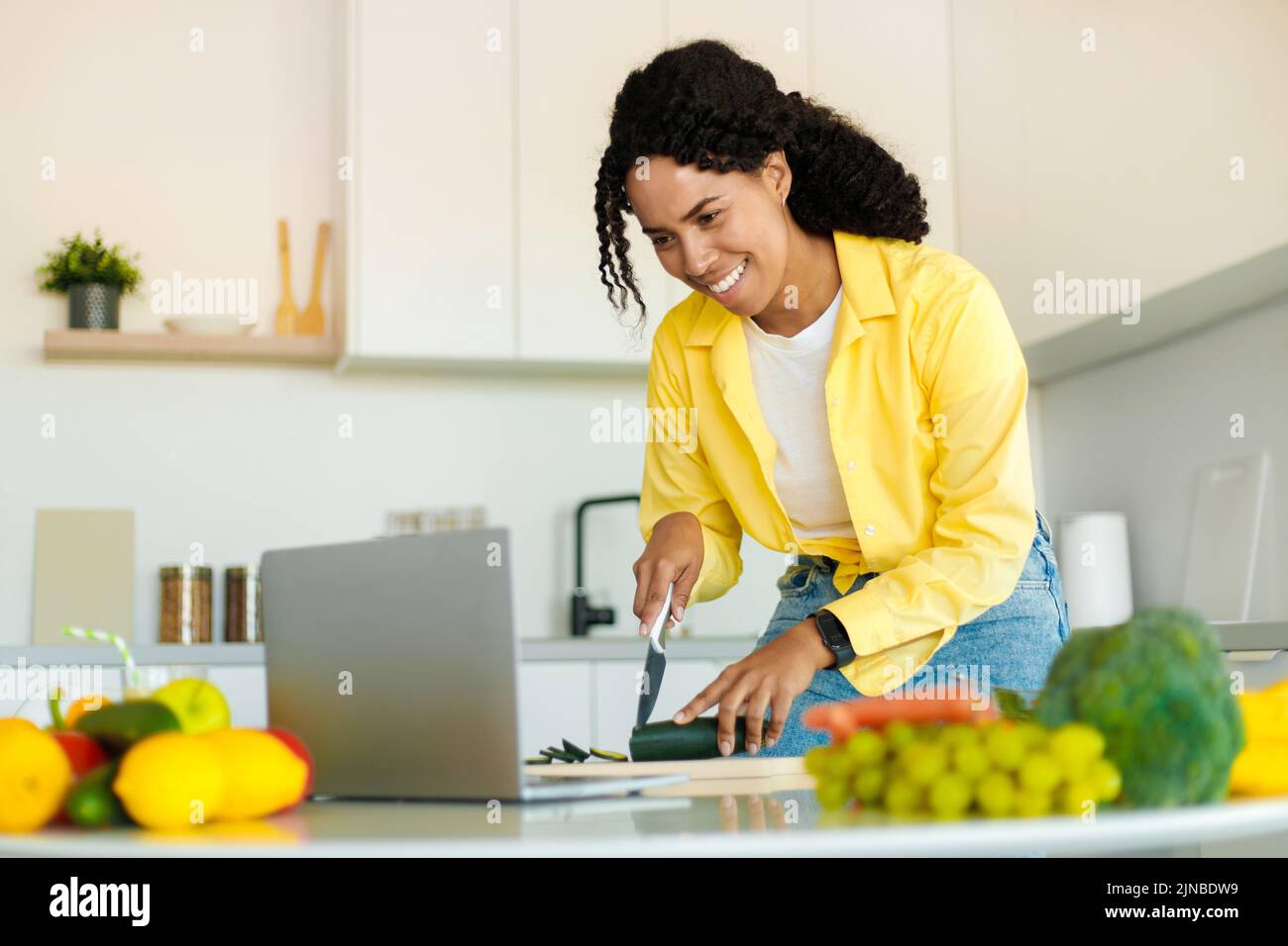 Nouvelle recette, blog alimentaire. Bonne jeune femme afro-américaine coupant des légumes dans la cuisine intérieure, en utilisant un ordinateur portable Banque D'Images