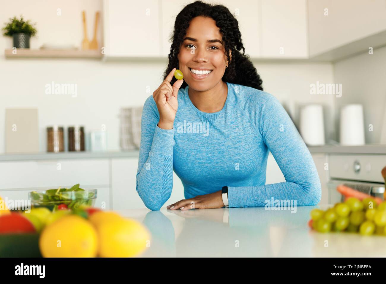 Femme afro-américaine excitée en forme de fruits pour le petit déjeuner, tenant le raisin et souriant à la caméra, assis dans la cuisine Banque D'Images