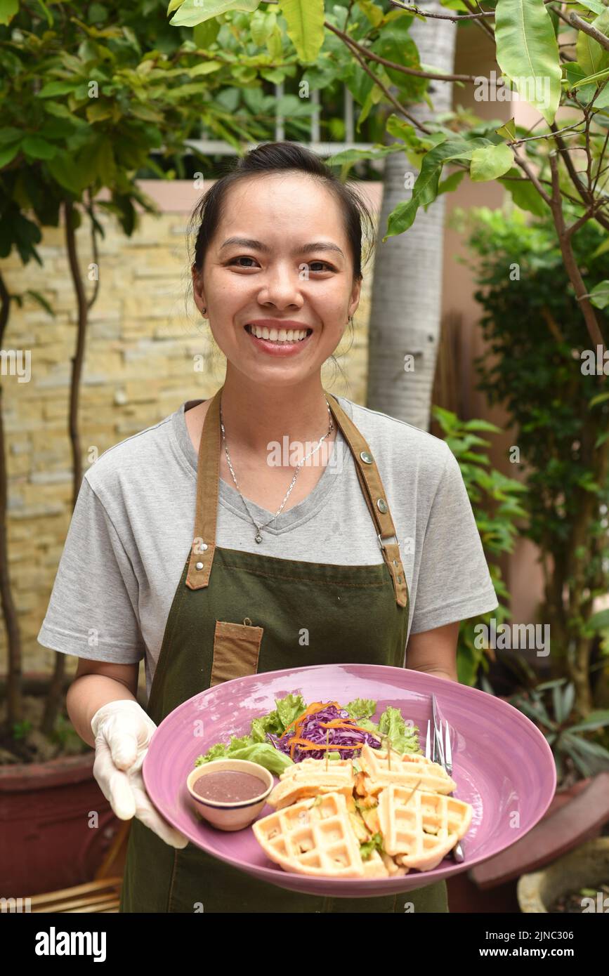 Une serveuse vietnamienne sert des gaufres belges avec des légumes verts et du poulet dans un café extérieur Banque D'Images