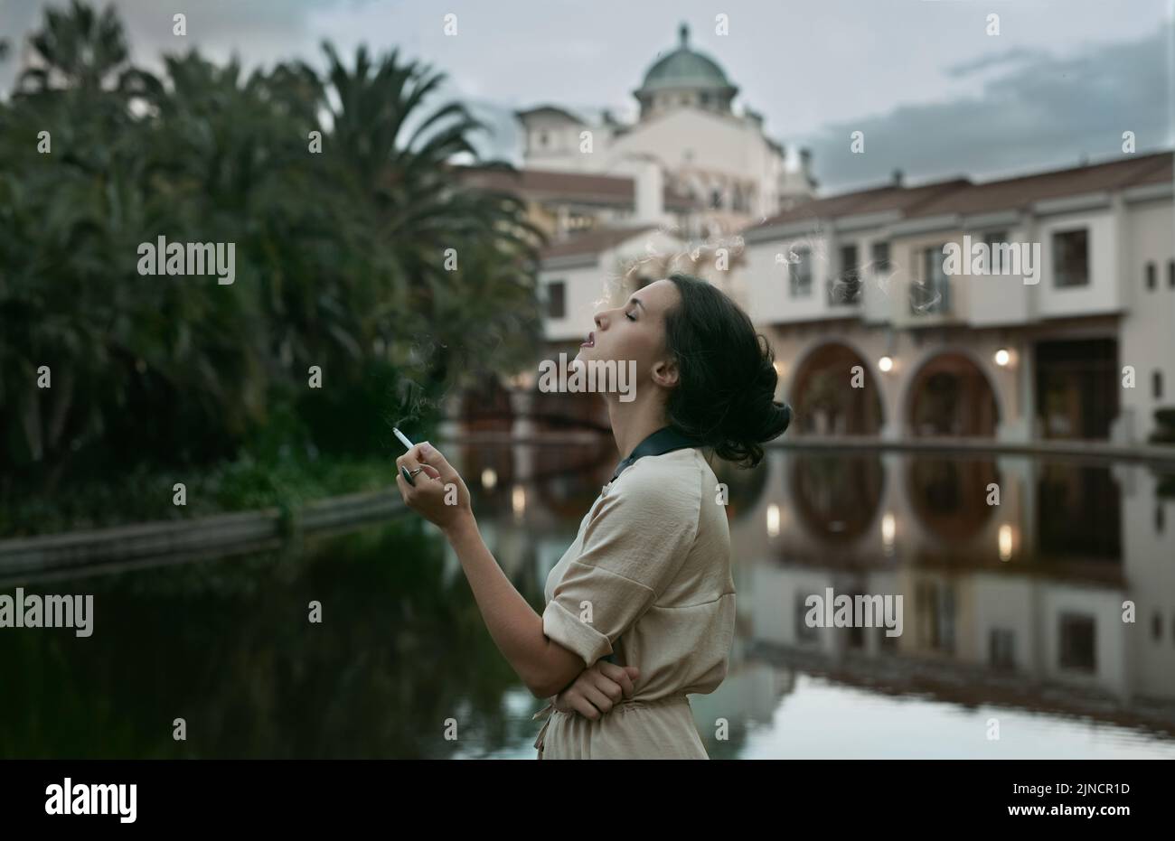La classe est intemporelle. Une jeune femme attrayante fumant une cigarette près d'un lac dans un cadre urbain. Banque D'Images