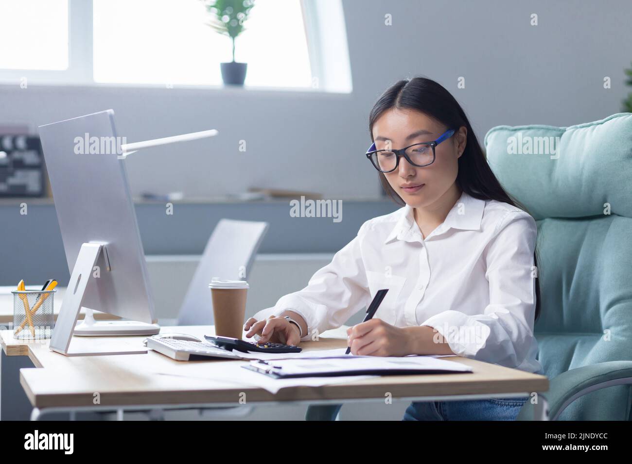 Workaholic. Une belle jeune femme d'affaires asiatique en lunettes est très tôt au travail, assis à la table buvant du café pour aller, une tasse de papier de café est debout sur le bureau près de l'ordinateur. Banque D'Images