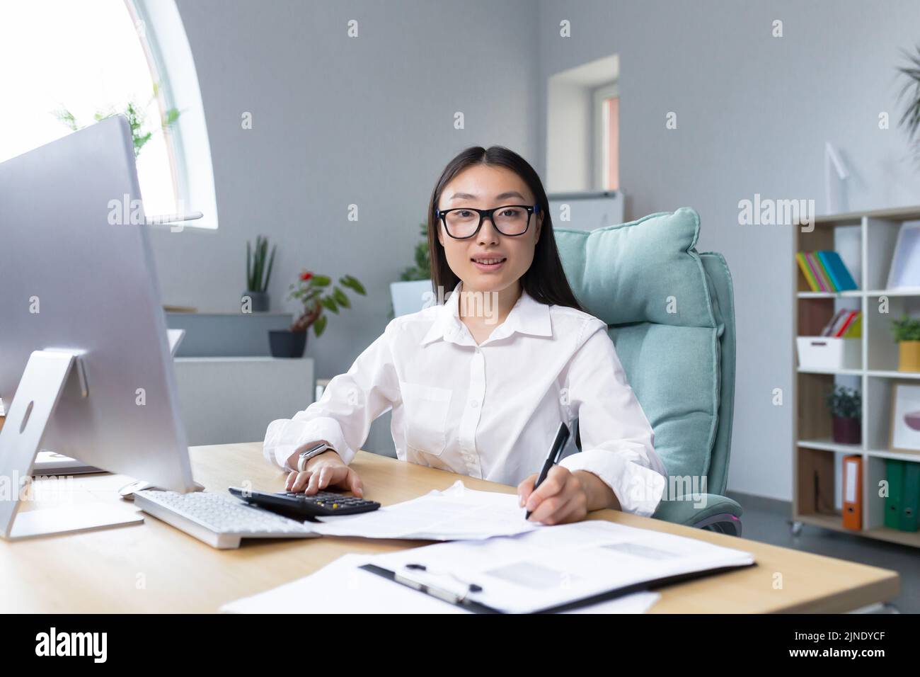 Travailler avec des documents. Portrait d'une jeune belle femme d'affaires asiatique comptable travaille avec des documents et des rapports. Assis au bureau, écrivant.il regarde la caméra, sourit Banque D'Images