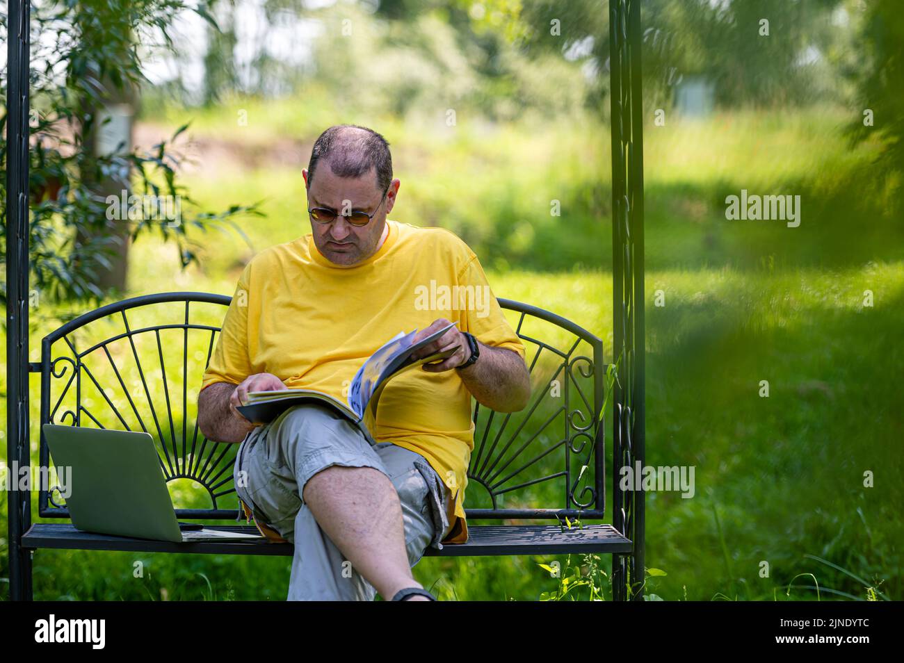 Homme mature avec ordinateur portable et documents travaillant à l'extérieur dans le jardin, concept de bureau à domicile vert. Banque D'Images