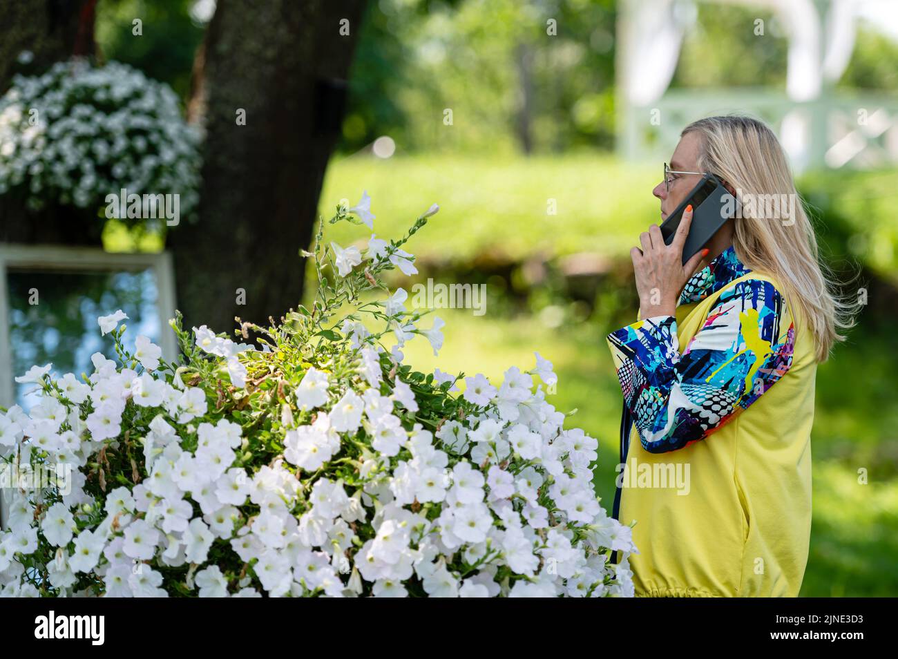 femme aîée blonde aux couleurs vives parlant sur smartphone dans le jardin, concept de communication en ligne Banque D'Images