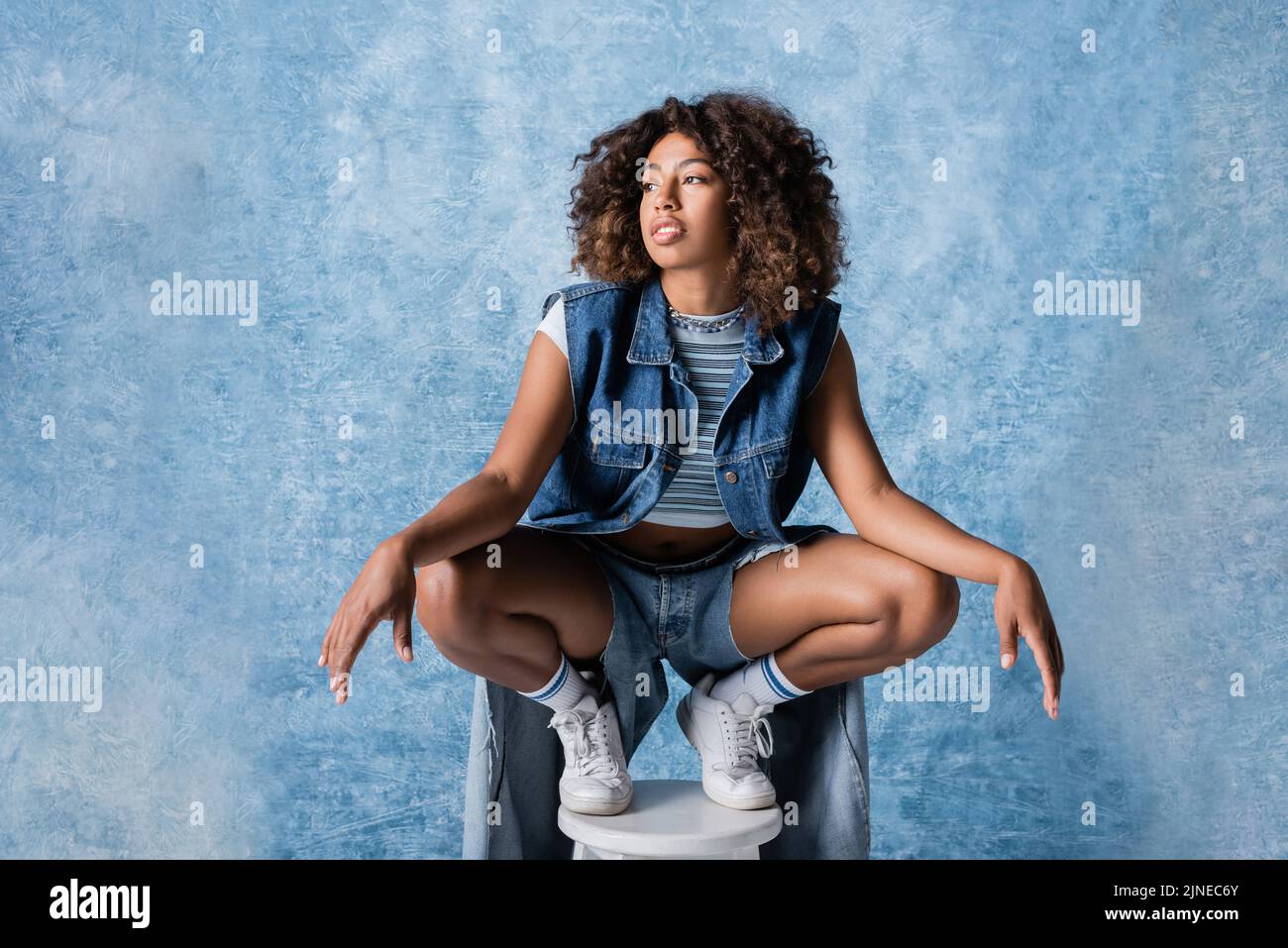 femme afro-américaine en vêtements denim tendance posé sur un tabouret sur fond bleu Banque D'Images