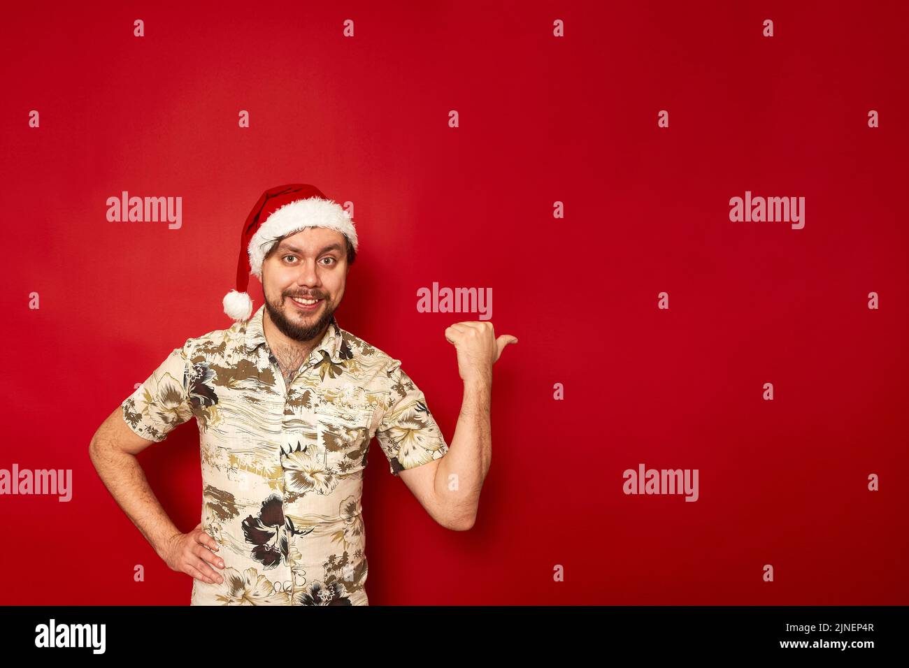 Portrait d'un homme souriant joyeux dans un chapeau de Noël et une chemise touristique pointe de son pouce à côté Banque D'Images